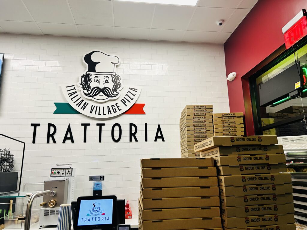 Image of a pizza shop. The wall is white subway tile, with a cartoonish chef’s face in black and white, with a red and green banner with the word “Trattoria” under it. Pizza boxes are stacked high.