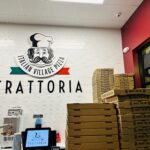 Image of a pizza shop. The wall is white subway tile, with a cartoonish chef’s face in black and white, with a red and green banner with the word “Trattoria” under it. Pizza boxes are stacked high.