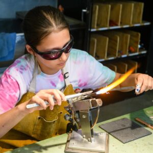 Photo shows a young girl working with a torch and a stick of glass to create art.